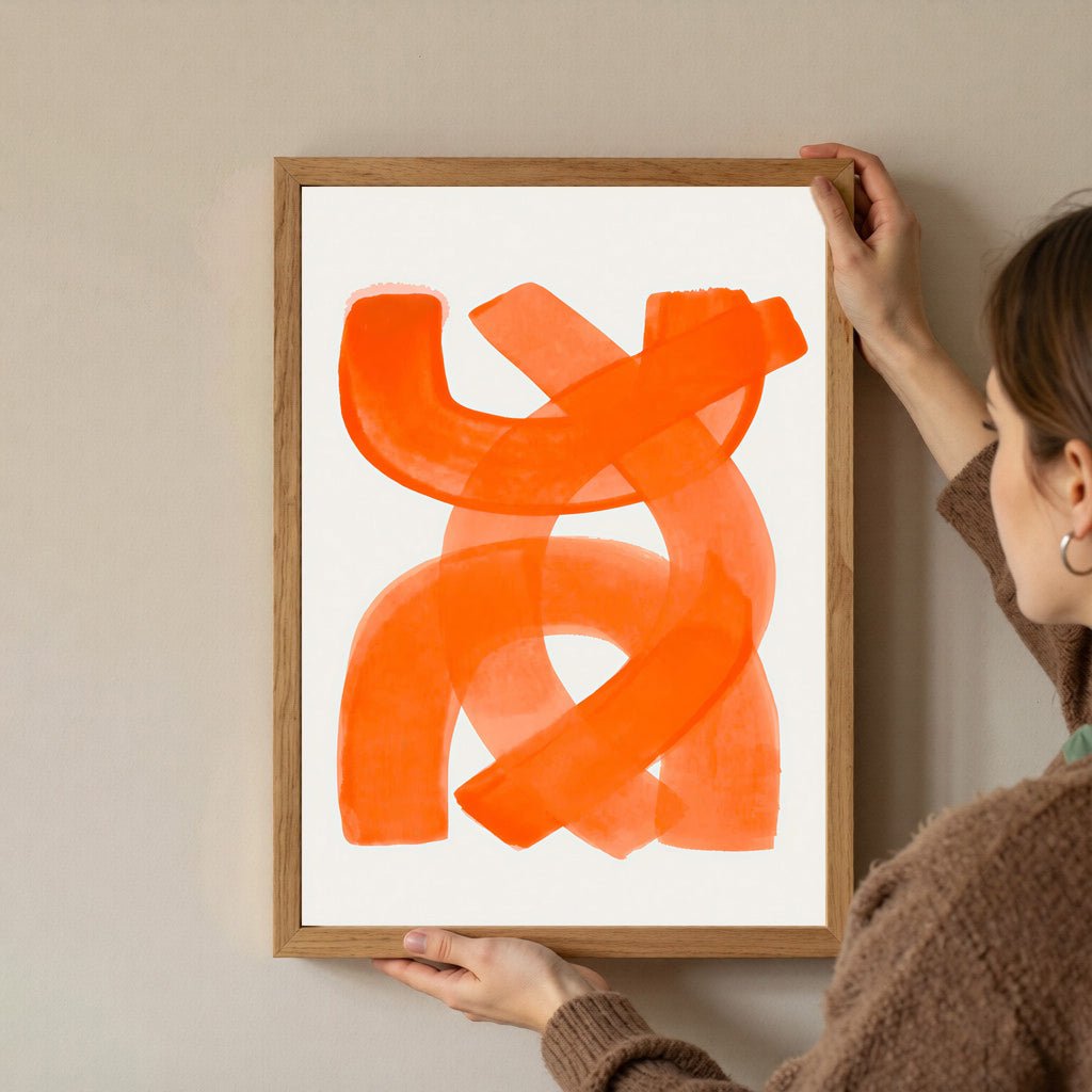 Person hanging a framed abstract orange artwork on a beige wall