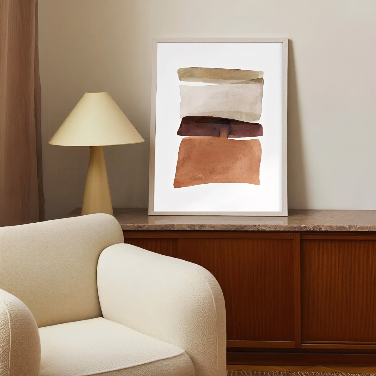 Framed abstract art on a wall above a wooden cabinet with a lamp and beige armchair.